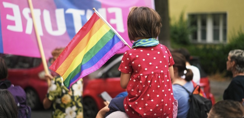 Mädchen auf der Schulter eines Erwachsenen mit Regenbogenfahne 