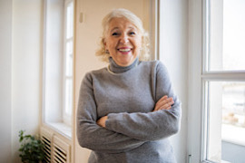 Ältere Dame mit verschränkten Armen lächelnd in ihrer Wohnung