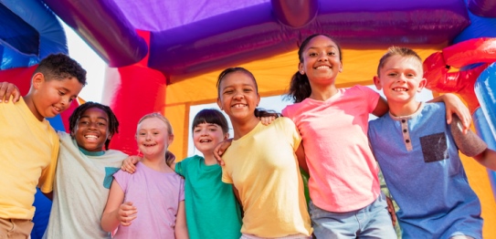 Eine Reihe von lachenden multiethnischen Kindern, die sich die Arme auf die schultern gelegt haben in einem bunten Hüpfhaus